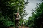 Ein Jäger steht mit Gewehr auf einem Waldweg, umgeben von dichter grüner Natur.