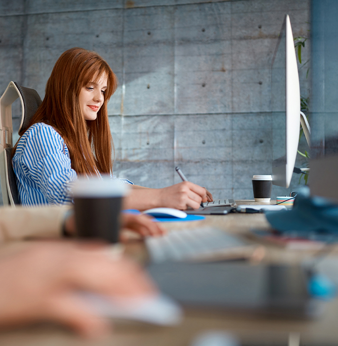 Smiling woman with red hair working at a desktop computer, taking notes in a bright office with coffee cups nearby.