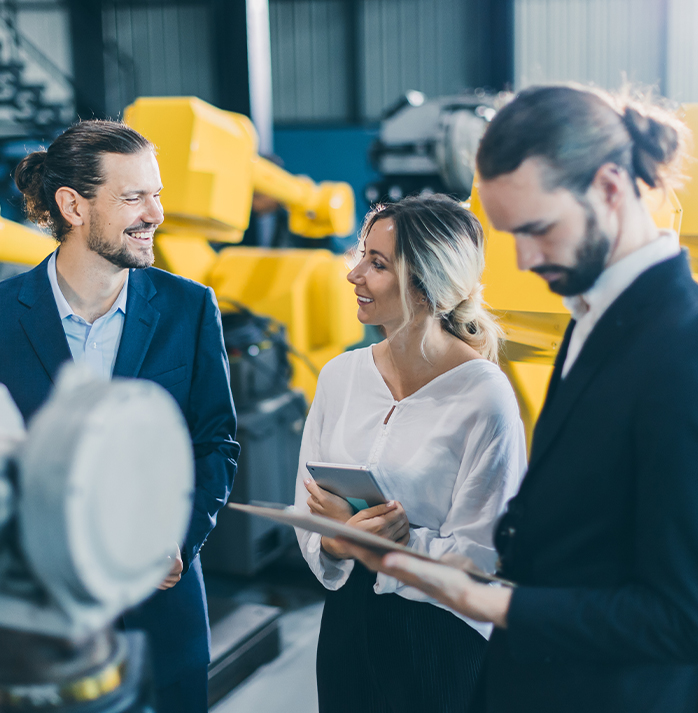 A customer and two sales men, standing in front of yellow industrial machinery, with one person holding a tablet